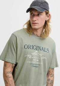 Oliven grøn bomulds t-shirt med teksten "ORIGINALS", hvide og marineblå grafik, afslappet pasform og korte ærmer, båret med en grå kasket.