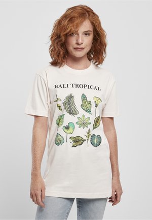 Femme aux cheveux roux portant un t-shirt écru "BALI TROPICAL" avec des illustrations variées de feuilles tropicales vertes, associé à un jean bleu clair.