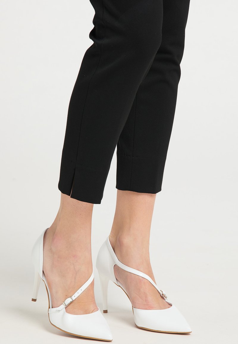 Scarpe bianche con tacco alto, dalla punta appuntita e una sottile cinghia alla caviglia. Materiale sintetico liscio, con una finitura lucida. Pantaloni neri fino alla caviglia.