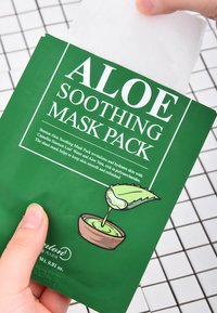 Packaging verde con testo in grassetto bianco che recita "MASCHERA LENITIVA ALL'ALOE". Include un'illustrazione di una foglia di aloe e una crema in una ciotola.
