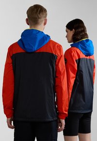 Chaqueta ligera en colores rojo, negro y azul, con capucha ajustable, puños elásticos y un parche de logo en la parte posterior.