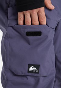 Pantalones impermeables morados con un bolsillo lateral, que cuentan con un cierre de Velcro negro y un parche de logo en la parte inferior.