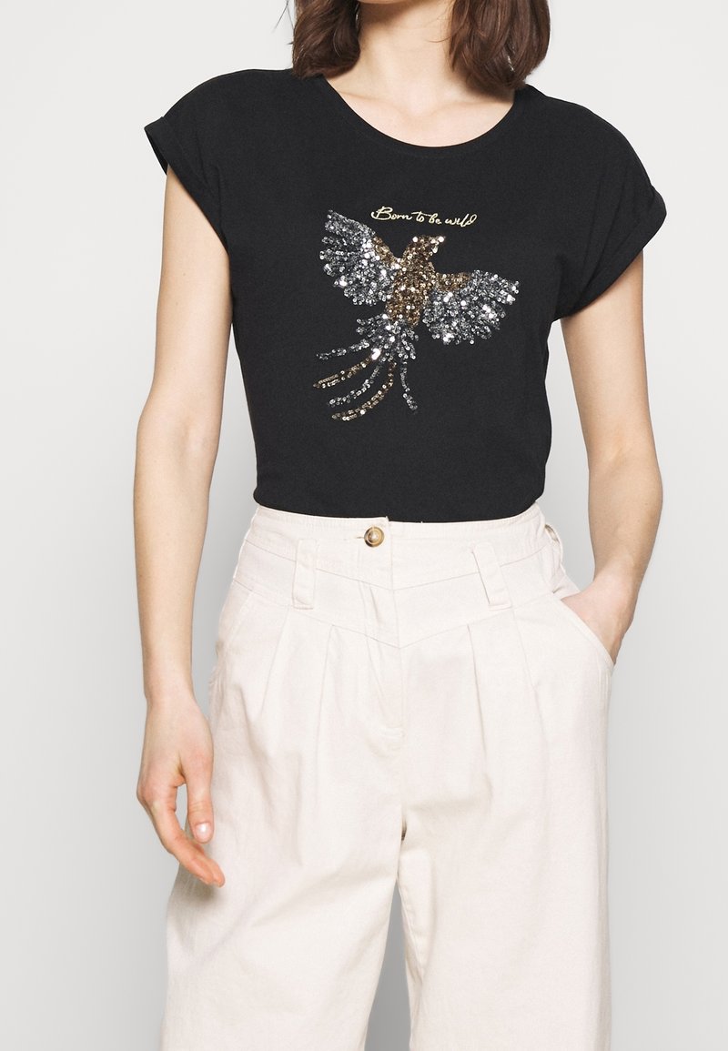 Femme portant un t-shirt noir avec un motif d'oiseau en sequins et le texte "Born to be wild", associé à un pantalon blanc taille haute.