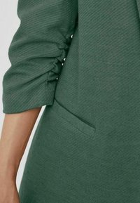 Groene textuurstof met een subtiel patroon, met gathered mouwen en een zijzak voor detail.