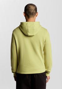 Tyngre sweatshirt i ljusgul, med huva och långa ärmar. Slät yta med avslappnad passform. Bakifrån med mörka byxor.