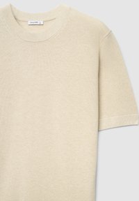 Haut en maille beige clair à manches courtes avec un col rond, présentant un motif tissé texturé et des poignets côtelés. L'étiquette affiche le nom de la marque "CALLIOPE".