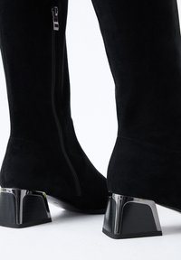 Las botas negras de ante hasta la rodilla cuentan con un zipper lateral y un tacón de cromo geométrico, combinando textura y elementos de diseño moderno.