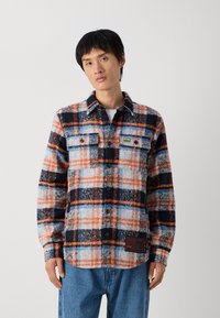 Camicia di flanella a quadri blu, arancione e bianca; presenta due tasche sul petto, chiusura con bottoni e tessuto testurizzato. Etichetta nera sul lato.