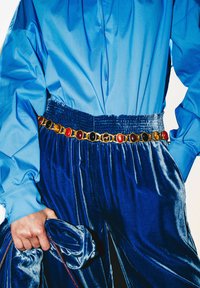 Blusa in raso blu con collo alto, abbinata a pantaloni in velluto blu scuro. Una cintura d'oro con pietre colorate aggiunge un dettaglio, sostenuta da un tessuto abbinato.