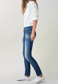 Jean skinny en denim bleu foncé, décoloré au niveau des genoux. Associé à une chemise blanche à manches longues et des baskets blanches basses, se tenant de profil.