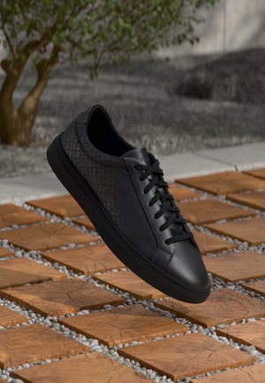 Sort lædersneakers med snørebånd, der svæver over træfliser og små grå sten udendørs med et træ i baggrunden.