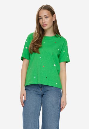 Donna con lunghi capelli castani che indossa una maglietta verde a maniche corte con piccoli puntini colorati e jeans blu, in piedi contro uno sfondo bianco.