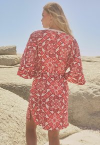 Robe portefeuille rouge et blanche à motifs avec des manches courtes, mettant en avant des motifs floraux et une taille cintrée. Portée devant un arrière-plan rocheux.