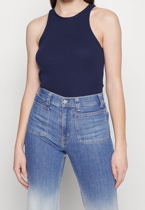 Kvinde iført en ærmeløs navyblå ribstrikket tanktop, stukket ned i højttaljede denimjeans i lyse til mørke nuancer med lynede frontlommer.