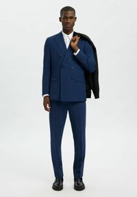 Abito blu a doppio petto con pantaloni coordinati, realizzato in un tessuto texturizzato. Camicia bianca sotto, giacca nera appoggiata sulla spalla, scarpe nere.