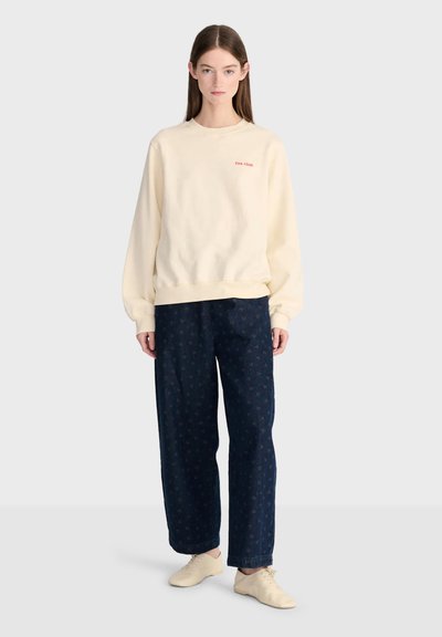 Femme aux longs cheveux bruns portant un sweat crème avec texte rouge, un pantalon large sombre avec un motif subtil, et des chaussures crème à lacets.