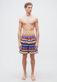 Άνδρας που στέκεται ξυπόλυτος φορώντας πολύχρωμα σορτς με σχέδιο ζιγκ ζαγκ, μπροστά σε ένα απλό λευκό φόντο, κοιτάζοντας στο πλάι.