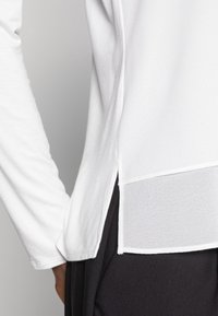 Haut blanc à manches longues avec un ourlet fendu en diagonale en tissu transparent en bas, associé à un pantalon sombre. Texture lisse.