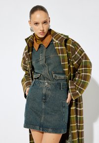 Jeansoverallklänning med fackfickor, bärs under en lång, rutig brun och grön kappa. Har en brun ribbad krage och tjockt, strukturerat tyg.