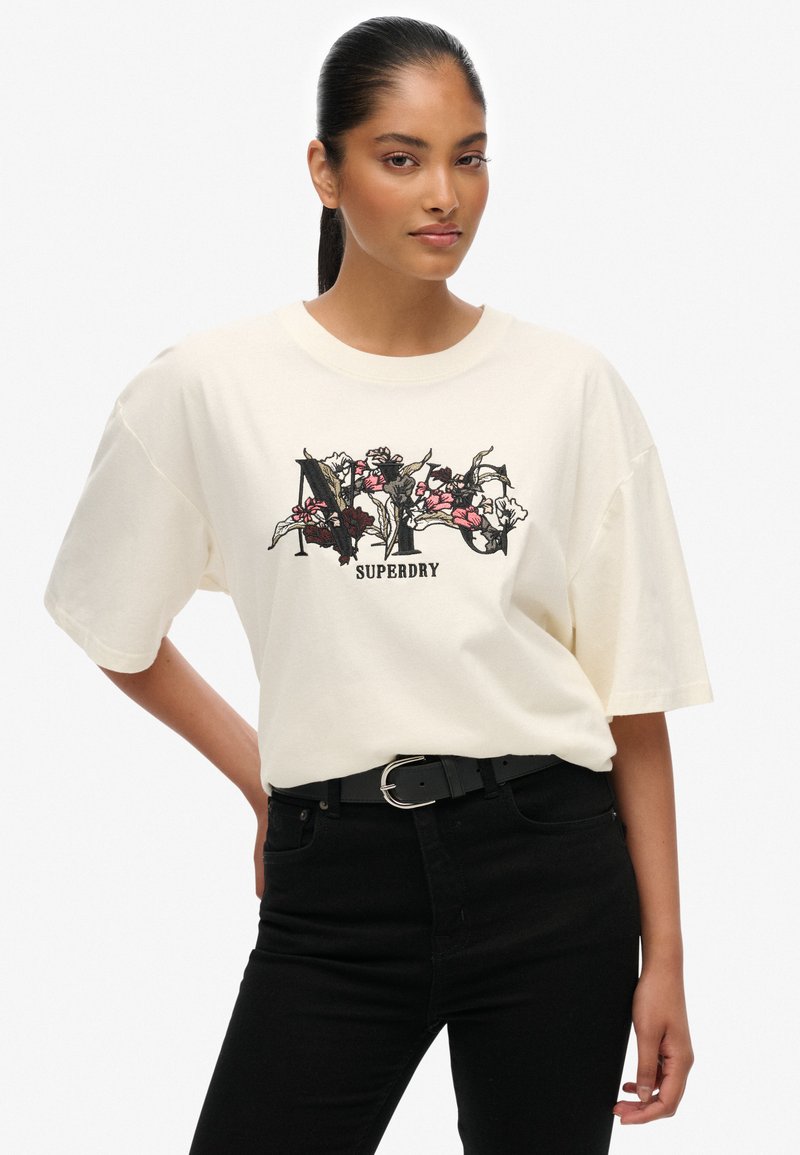 T-shirt in cotone crema con design grafico floreale e testo "SUPERDRY". Vestibilità ampia, colletto rotondo, maniche corte. Abbinato a pantaloni neri e cintura.