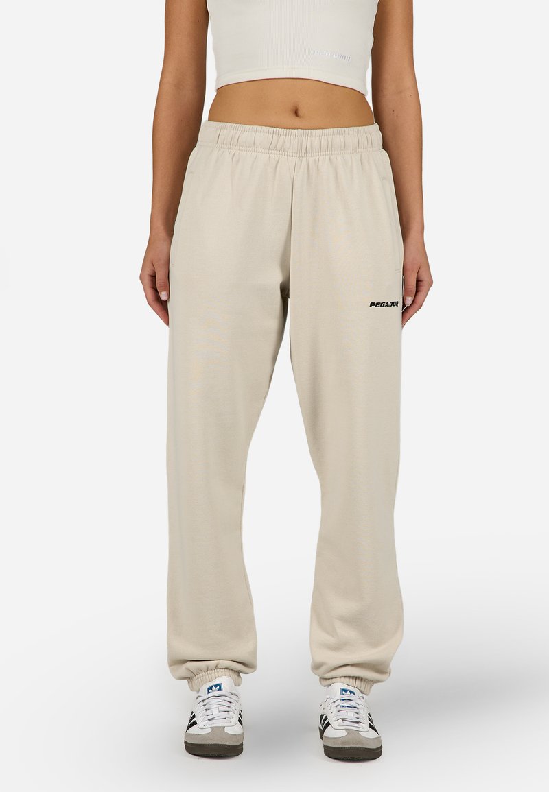 Béžové tepláky s elastickým pásom a manžetami, s voľným strihom a malým čiernym logom na ľavom stehne. Mäkkučký textil.