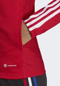 Red athletic shirt featuring a textured mesh panel, white triple stripes on the sleeve, and an "AEROREADY" logo at the hem.