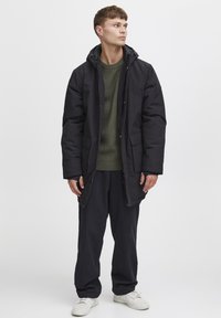 Parka aislante negra con capucha, que presenta un cierre de cremallera y botones a presión, bolsillos laterales, llevada sobre un suéter de canalé verde oscuro y pantalones oscuros.