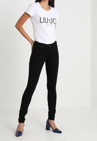 Camiseta blanca con estampado "LIUJO MILANO", combinada con jeans negros ajustados. La modelo lleva tacones azules de punta. Diseño simple y limpio, silueta ajustada.