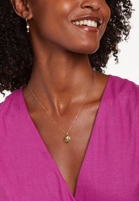 Gouden ketting met een ronde hanger met een gestructureerd ontwerp, gedragen boven een diep roze halslijn. Oorbellen met kleine kralen zijn zichtbaar.