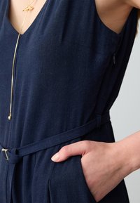Tuta navy con scollatura a V, vita cintata e tasche. Realizzata in tessuto testurizzato, presenta una leggera lucentezza e un design minimalista.