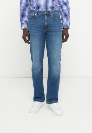 Mann trägt blaue Jeans, ein blau-weiß kariertes Hemd und weiße Sneakers, steht vor weißem Hintergrund.