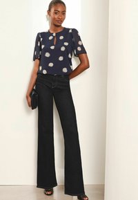 Marinebluse med hvide prikker, korte ærmer, nøglehuludskæring; parret med sorte flared jeans og sort clutch. Glat stof, moderne snit.