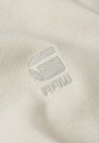 Broderad logotyp på ljusbeige tyg, som visar en subtil textur med en minimalistisk design och ett mjukt, slätt utseende.