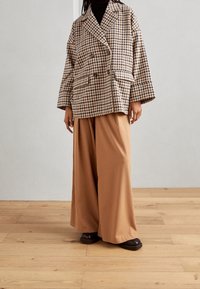 Blazer doppiopetto in un motivo houndstooth marrone e crema, abbinato a pantaloni larghi beige. Presenta grandi tasche e una vestibilità comoda.