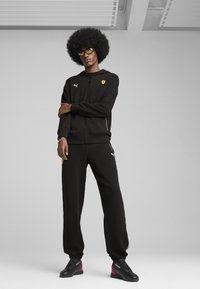 Sort zip-up hættetrøje og joggingbukser, begge med et gult Ferrari-emblem. Tekstureret stof, slim fit, med kontrasterende lynlås og slanke sorte sko.