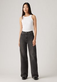 Un débardeur blanc à côtes associé à un jean en denim noir à taille haute et jambes larges. Texture lisse avec un design classique à cinq poches.