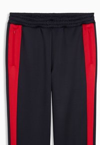 Φόρμα παντελόνι σε ναυτικό μπλε με κόκκινες λωρίδες στα πλάγια, διαθέτει ελαστική μέση και πλαϊνές τσέπες με φερμουάρ για λειτουργικότητα. Ομαλή υφή υφάσματος.