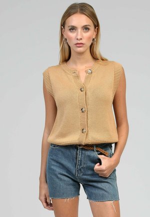 Femme portant un gilet tricoté beige sans manches boutonné et un short en jean bleu avec ceinture tressée, debout devant un fond gris uni.