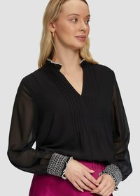 Schwarze Bluse mit transparenten langen Ärmeln, plissiertem Vorderteil und gemusterten Manschetten. Besitzt einen V-Ausschnitt und kontrastierende Besätze am Kragen.