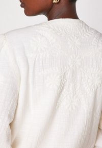 Persona di carnagione scura che indossa una blusa bianca con texture e ricami floreali sulla schiena, accompagnata da un grande orecchino rotondo d'oro.