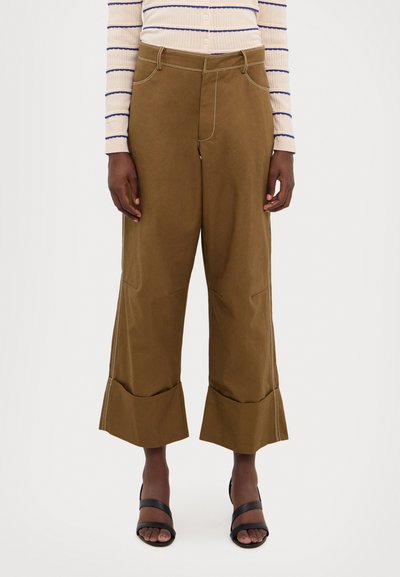 Pantalones marrones de pierna ancha con dobladillo acampanado, costuras en contraste y bolsillos laterales, combinados con sandalias negras.