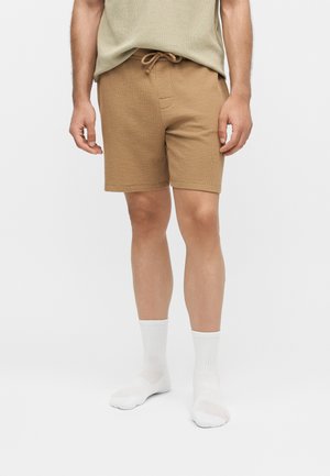 Férfi, aki bézs színű kötött rövidnadrágot visel húzózsinórral, bézs felsővel és fehér bokazoknival, áll egy egyszerű világos háttér előtt.