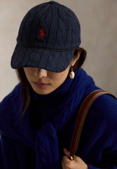 Marineblå strikket lue med kabelmønster og et rødt emblem på fronten; kombinert med en blå strikket genser og brun skulderveskerem.