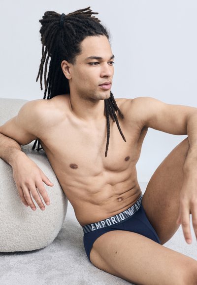 Άνδρας χωρίς πουκάμισο με μακριά πλεξούδες κάθεται στο πάτωμα, στηριζόμενος σε μαξιλάρι κρεμ υφής, φορώντας σκούρα μπλε εσώρουχα Emporio Armani.