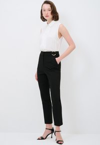 Witte mouwloze blouse met een knopenlijst, gecombineerd met hoge zwarte broeken met een gouden ronde riemdetail en hoge sandalen.