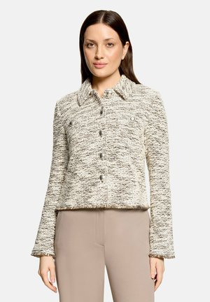 Femme portant une veste texturée crème et marron boutonnée avec un col et un pantalon beige, debout devant un fond clair uni.