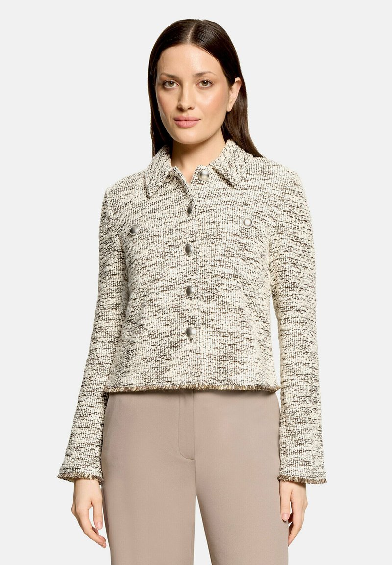 Femme portant une veste texturée crème et marron boutonnée avec un col et un pantalon beige, debout devant un fond clair uni.