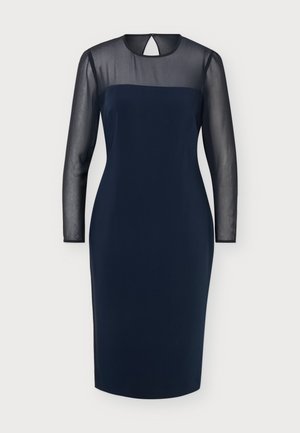 Femme debout portant une robe bleu marine longueur genoux avec des manches longues transparentes et une partie supérieure de la poitrine, associée à des chaussures plates noires.