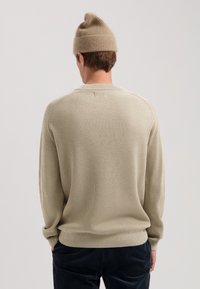 Beiger gerippter Strickpullover mit langen Ärmeln und rundem Halsausschnitt, kombiniert mit einer passenden, hellen Strickmütze. Dunkle Hose sichtbar.