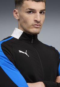 Crna sportska jakna s plavim detaljima i bijelim ovratnikom, koja sadrži zatvaranje na patentni zatvarač i istaknuti bijeli Puma logo na lijevoj strani prsnog koša.